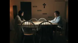 A man and woman sitting across the dining room table from one another, a cross hung on the wall.